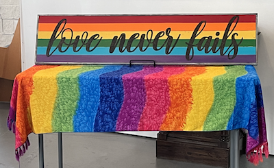 Table with pride rainbow cloth and sign saying "Love never fails" on top of it